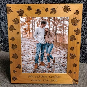 May include: A wooden picture frame with a carved autumn leaf design. The frame holds a photo of a couple standing in a wooded area. The frame is engraved with the text "Mr. and Mrs. Lindner October 17th, 2020".