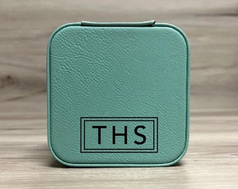 May include: A teal blue square-shaped jewelry box with a black leather-like texture. The box has a black border around the letters "THS" in the center.