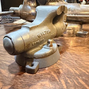May include: A grey metal Wilton bench vise with a swivel base and a cylindrical handle, featuring the embossed text Wilton USA on the side. The tool has a textured finish, a circular mounting plate with bolt holes, and a metallic screw mechanism.