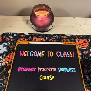 May include: A black digital drawing tablet with a yellow border, displaying the text "WELCOME TO CLASS! Beginner Procreate Seamless Course" in colorful letters. The tablet is on a black and white Halloween-themed mousepad with a lit candle in the background.