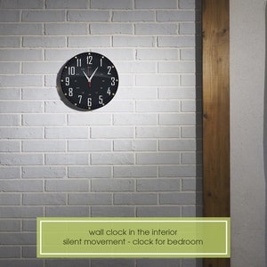 May include: A black wall clock with white numbers and gold accents hangs on a white brick wall. The clock has a silent movement and is ideal for a bedroom.