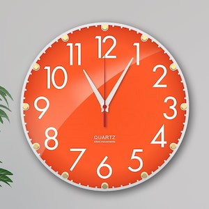 Può includere: Orologio da parete arancione con numeri e lancette bianchi. L'orologio ha un bordo bianco e dettagli dorati. Il quadrante dell'orologio dice "QUARTZ silent movement".