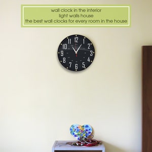 May include: A black wall clock with white numbers and gold accents hangs on a light beige wall. The clock is in a room with a wooden door and a small table with a decorative heart.