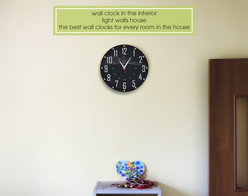 May include: A black wall clock with white numbers and gold accents hangs on a light beige wall. The clock is in a room with a wooden door and a small table with a decorative heart.