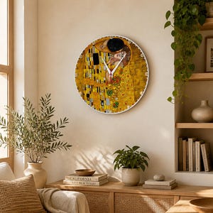 Puede incluir: Un reloj de pared redondo con la obra "El Beso" de Gustav Klimt. La esfera del reloj muestra las icónicas imágenes doradas y coloridas de la pareja abrazándose. El reloj tiene un marco blanco y está montado en una pared beige, agregando un toque artístico a la habitación.