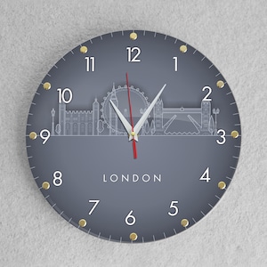 May include: A gray round clock with gold accents and white numbers. The clock face features a silhouette of the London skyline, including Big Ben, the London Eye, and Tower Bridge. The clock face also includes the word "LONDON" in white text.