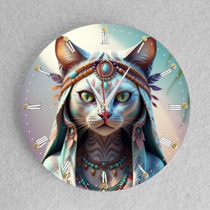 Op de afbeelding: Een ronde klok met Romeinse cijfers heeft een kleurrijke illustratie van een kat die een hoofdtooi draagt met veren en turquoise juwelen. De ogen van de kat zijn groen en de achtergrond is een verloop van blauw naar roze.