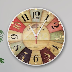 Puede incluir: Un reloj redondo con marco blanco y una cara de estilo madera. La cara tiene un diseño colorido con números romanos y el texto "CAE de la TOUR" y "PARIS FRANCE".