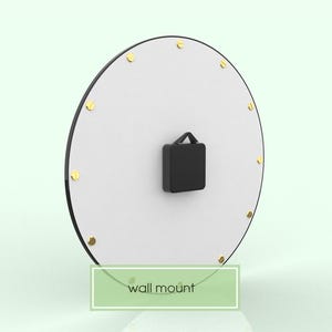 以下が含まれることがあります： 黒の縁取りと金のアクセントが付いた白い丸い時計。時計は壁掛け式で、黒い文字盤があります。