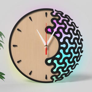 Puede incluir: Un reloj de pared redondo con una esfera de madera clara y borde negro. El reloj presenta marcadores de hora negros y manecillas blancas. Una sección del reloj tiene un diseño negro ondulado con un degradado de colores rosa, azul y verde.