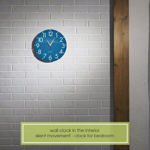 May include: A blue wall clock with white numbers and hands against a white brick wall. The clock has the text "silent movement - clock for bedroom" below it.