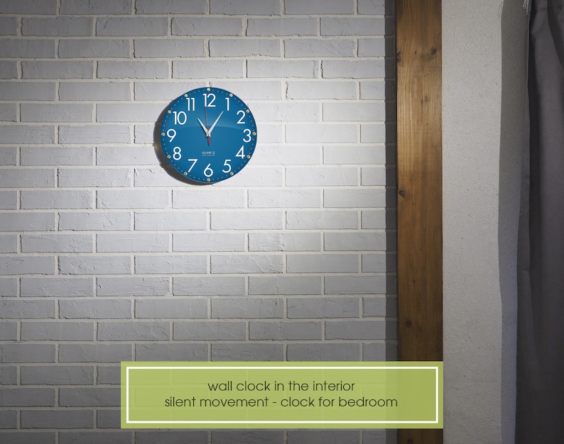 May include: A blue wall clock with white numbers and hands against a white brick wall. The clock has the text "silent movement - clock for bedroom" below it.