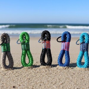 Può includere: Cinque portachiavi colorati in corda con anelli metallici sono esposti su una spiaggia sabbiosa con l'oceano sullo sfondo. I portachiavi sono grigi, verdi, neri, blu e turchese, ognuno con una fascia a contrasto.