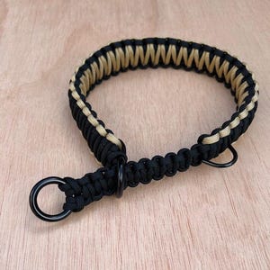 May include: A black and tan paracord dog collar with black metal rings and hardware. The collar is made of woven nylon cord. The collar is displayed on a light brown wooden surface.