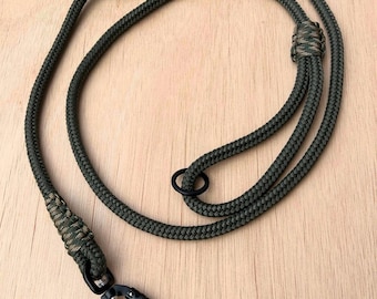 May include: A dark green lanyard made of braided rope, featuring a black carabiner clip and a black metal ring. The lanyard is designed for attaching keys, ID badges, or other small items. The background is a light wood surface.