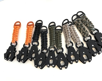 Puede incluir: Una colección de llaveros de paracord en varios colores, incluyendo naranja, negro, verde y camuflaje. Cada llavero tiene un cierre de metal negro y un anillo en D. Se muestran sobre un fondo blanco.