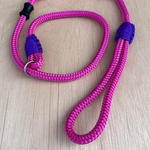 May include: A vibrant pink slip lead dog leash with purple detailing. The leash is constructed from a thick, braided rope, featuring a loop handle and a metal ring. A black plastic piece secures the loop.