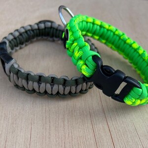 May include: Two paracord bracelets. One is green, gray, and tan, and the other is bright green and yellow. Both bracelets have black plastic buckles and metal rings. The bracelets are on a light wood surface.