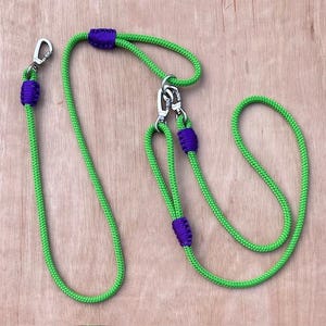 May include: A vibrant green dog lead with purple detailing. The lead is constructed from a robust rope material and includes silver metal clasps and connectors. It features a loop handle and a separate loop for attaching to a dog's collar.