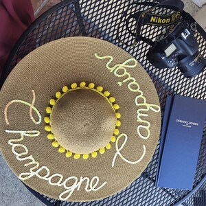 May include: A brown straw sun hat with yellow pom-poms around the brim. The hat has the words "Champagne and Prosecco" written on it in white.