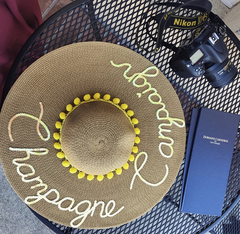 May include: A brown straw sun hat with yellow pom-poms around the brim. The hat has the words "Champagne and Prosecco" written on it in white.