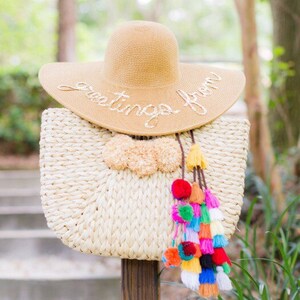 May include: A straw beach bag with colorful pom-poms hanging from the handle. A straw sun hat with the text "Greetings from" is sitting on top of the bag.