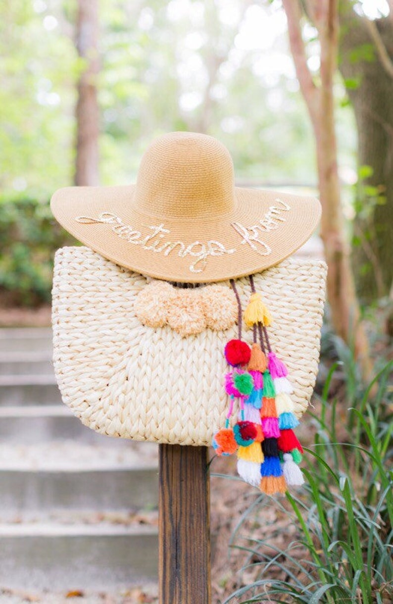 May include: A straw beach bag with colorful pom-poms hanging from the handle. A straw sun hat with the text "Greetings from" is sitting on top of the bag.
