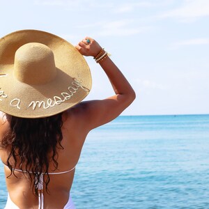 May include: A woman wearing a straw sun hat with the text "Leave a message" embroidered on it. She is standing on a beach with the ocean in the background.