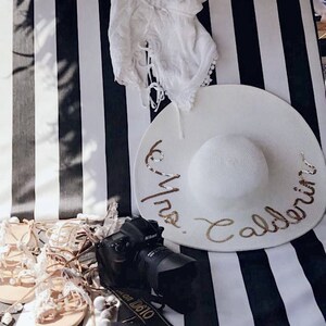 May include: White and black striped beach chair with a white sun hat with gold lettering that says "Mrs. Caldeira". There is a white lace top, a camera, and sandals on the chair.