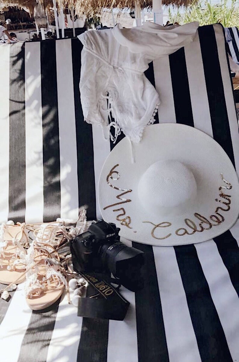 May include: White and black striped beach chair with a white sun hat with gold lettering that says "Mrs. Caldeira". There is a white lace top, a camera, and sandals on the chair.