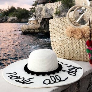 May include: White straw sun hat with black trim and the text "rest all day" written in black script. A woven straw tote bag with colorful tassels and pom-poms.