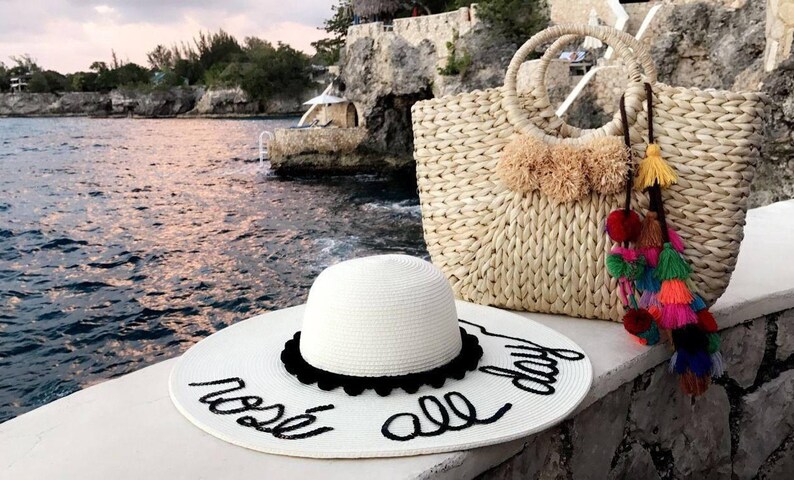 May include: White straw sun hat with black trim and the text "rest all day" written in black script. A woven straw tote bag with colorful tassels and pom-poms.