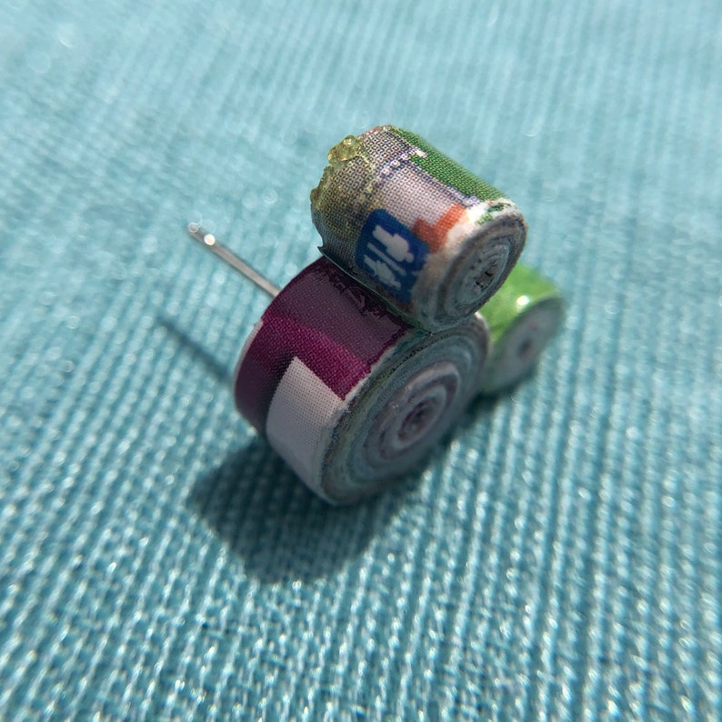 May include: A pair of earrings made from rolled up paper with a purple and green color scheme. The earrings are decorated with a pattern of people and a green and white pattern.