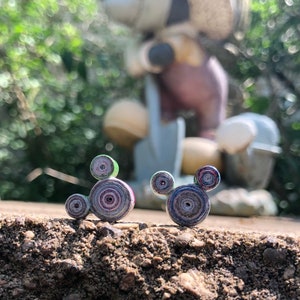 May include: Two small, colorful, paper-rolled Mickey Mouse figures. The figures are made of rolled paper with a variety of colors, including green, purple, and blue. The figures are sitting on a brown, dirt-like surface.