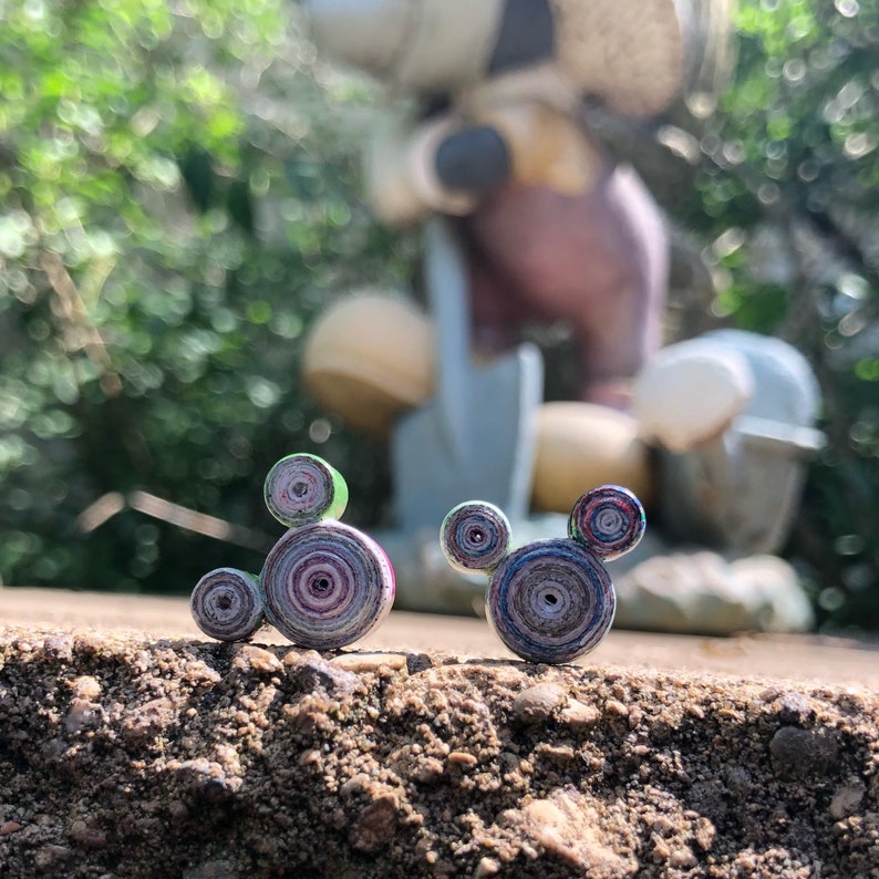 May include: Two small, colorful, paper-rolled Mickey Mouse figures. The figures are made of rolled paper with a variety of colors, including green, purple, and blue. The figures are sitting on a brown, dirt-like surface.