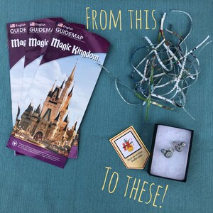 May include: A close-up of a small box with a pair of Mickey Mouse ears made from shredded paper. The box is sitting on a blue surface with a yellow sign that says "Find us on Etsy BaffeCrafte". There are three stacks of folded paper maps with the text "English Guidemap Magic Kingdom" on the front.