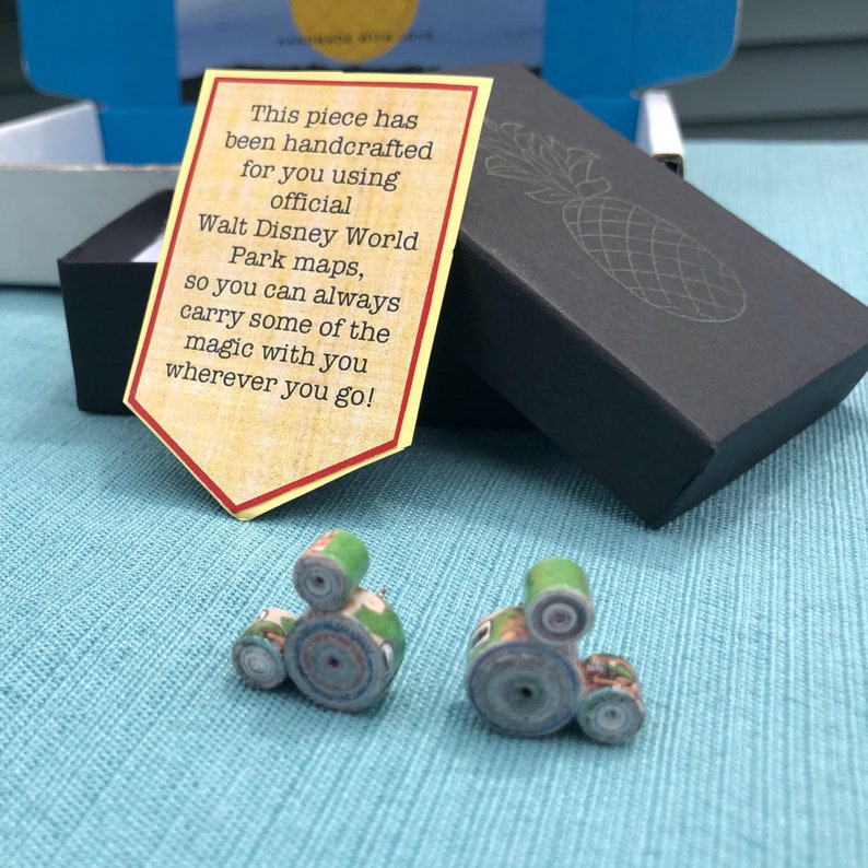 May include: A pair of green and blue paper-rolled earrings shaped like the iconic Mickey Mouse head. The earrings are packaged in a black box with a white pineapple design and a note that reads "This piece has been handcrafted for you using official Walt Disney World Park maps, so you can always carry some of the magic with you wherever you go!"