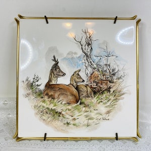 May include: Square ceramic tile with a hand-painted illustration of three deer resting in a grassy field, framed by a gold-toned metal frame. The artwork features a detailed depiction of the deer and their natural habitat.