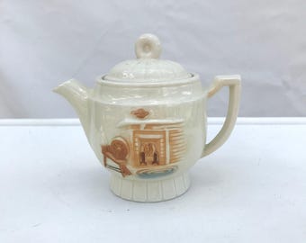 May include: A white ceramic teapot with a brown and orange design of a cabin with a fireplace. The teapot has a lid with a knob on top and a spout on the side.