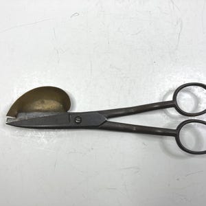 Puede incluir: Tijeras metálicas antiguas con protector de latón. Las tijeras tienen un aspecto oscuro y envejecido, con una hoja curva y asas de anillo. El protector de latón está unido a la hoja cerca del filo. Es probable que las tijeras sean para una artesanía o un oficio específico.