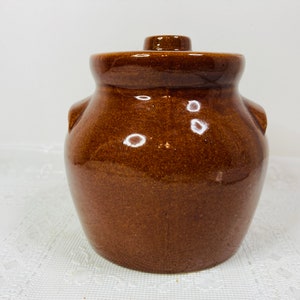 May include: A brown ceramic jar with a lid. The jar has a rounded body with two small handles and a lid with a small knob. The jar is a medium brown color and appears to be glazed.