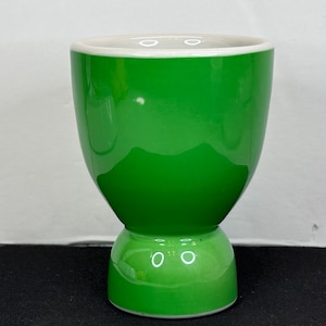 May include: A glossy green ceramic egg cup with a white rim and a pedestal base. The egg cup has a smooth, rounded shape and is designed to hold a single egg. The bright green color gives it a fresh, modern look.