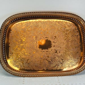 May include: A gold-colored, rectangular serving tray with ornate, embossed floral and scrollwork patterns. The tray has a decorative, rope-like border and a pierced edge. The tray is likely made of metal.
