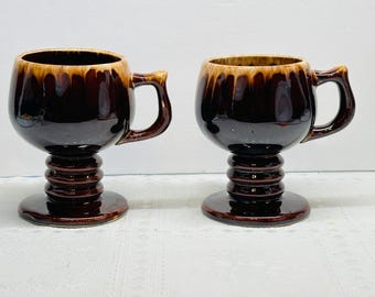 May include: Two matching brown ceramic mugs with a unique design. Each mug has a bulbous body, a handle, and a pedestal base. The interior of the mugs has a lighter brown color. The mugs are likely vintage or retro-style drinkware.