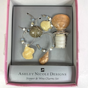 May include: A set of wine accessories in a pink box. The set includes a bottle stopper with a seashell design and several wine charms with seashell pendants. The box reads "Ashley Nicole Designs Stopper & Wine Charms Set."