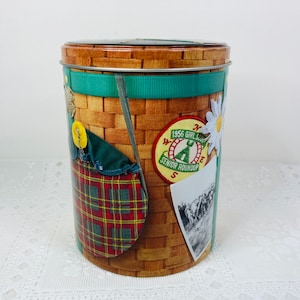 May include: A brown and green tin container with a wicker basket design. The container has a green ribbon and a variety of patches, including a Girl Scout Senior Roundup patch from 1956.