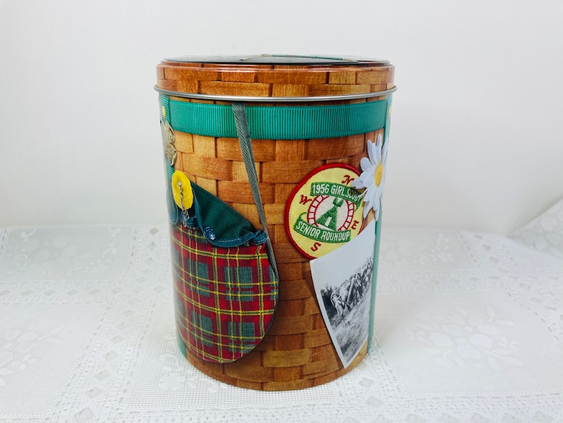 May include: A brown and green tin container with a wicker basket design. The container has a green ribbon and a variety of patches, including a Girl Scout Senior Roundup patch from 1956.