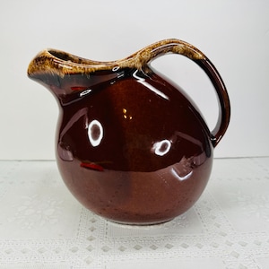 May include: A glossy, dark brown ceramic pitcher with a rounded body, a curved handle, and a wide pouring spout. The rim features a decorative drip glaze effect in a lighter tan color, contrasting with the deep brown finish of the main vessel.