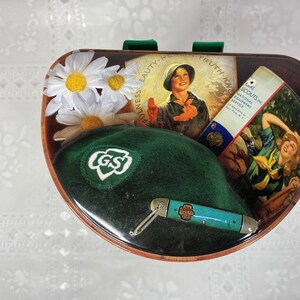 May include: A green Girl Scout hat with a white GS logo, a small pocket knife, and a vintage Girl Scout pamphlet with the text "Girl Scouts National Equipment Service" on a brown background with white daisies.