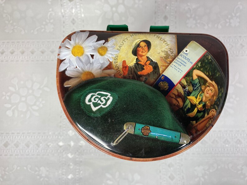 May include: A green Girl Scout hat with a white GS logo, a small pocket knife, and a vintage Girl Scout pamphlet with the text "Girl Scouts National Equipment Service" on a brown background with white daisies.